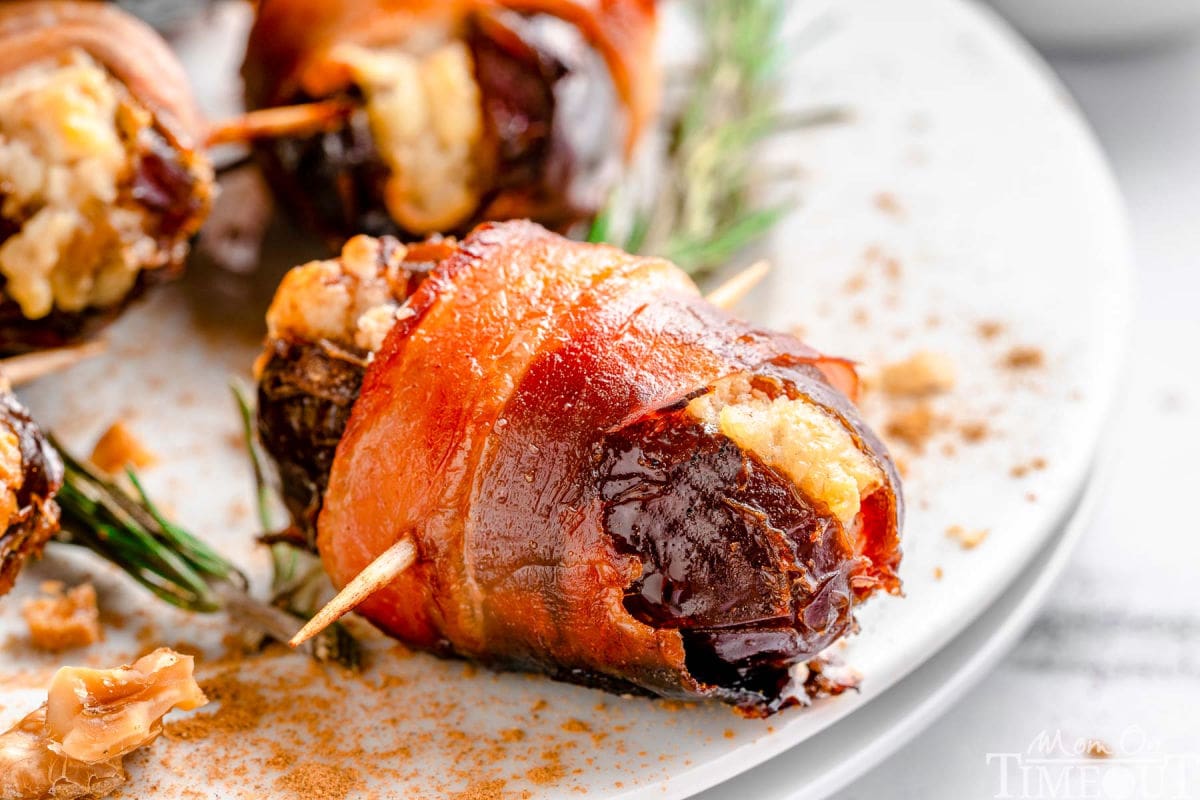 Wide shot of dates wrapped in bacon served on round white plate garnished with rosemary.