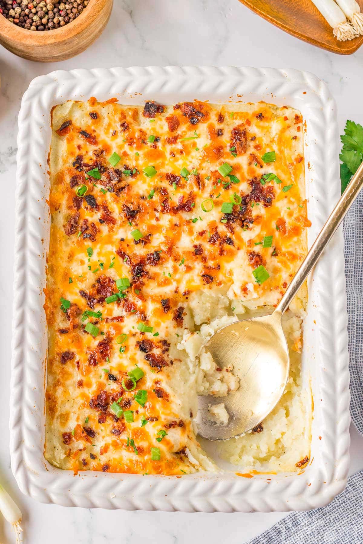 Top down look at loaded mashed potato casserole in a white baking dish with a gold serving spoon resting in the dish where a serving has been removed.