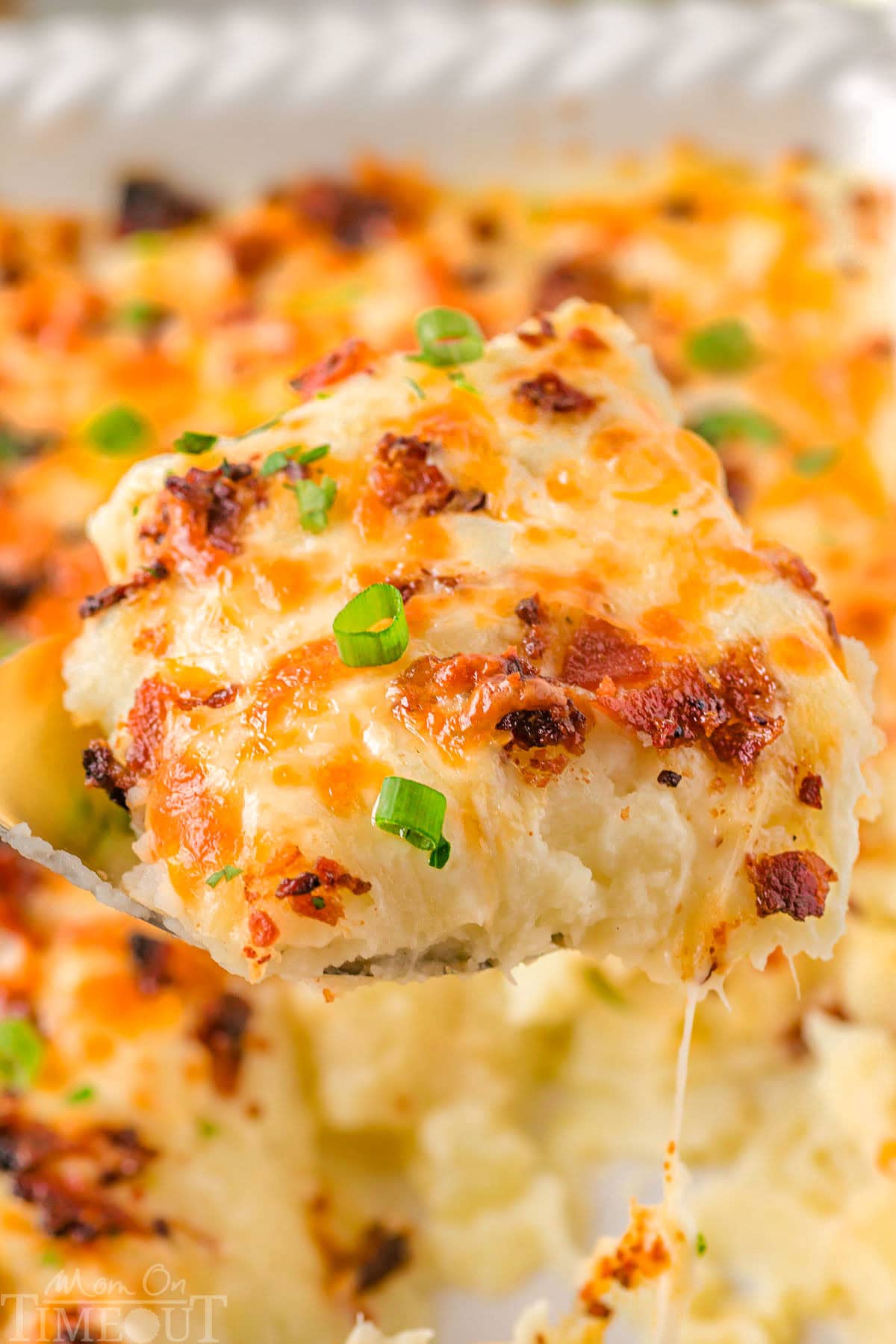 Serving of mashed potato casserole being lifted out of a white baking dish with the rest of the casserole in it.