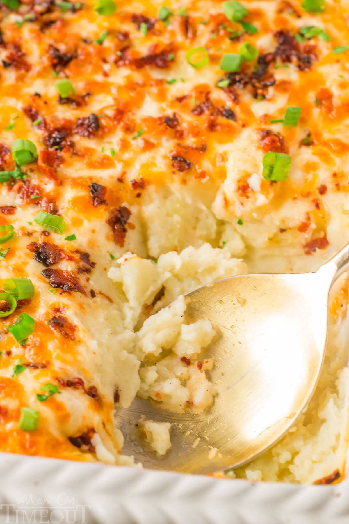 Top down look at loaded mashed potato casserole recipe in a white baking dish with a gold serving spoon resting in the dish where a serving has been removed.