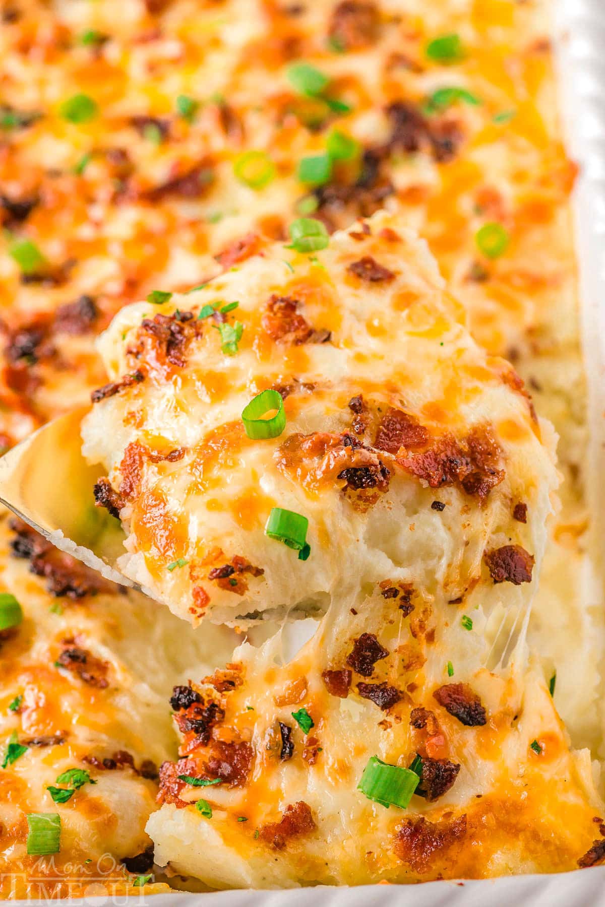 Serving of mashed potato casserole being lifted out of a white baking dish with the rest of the casserole in it.