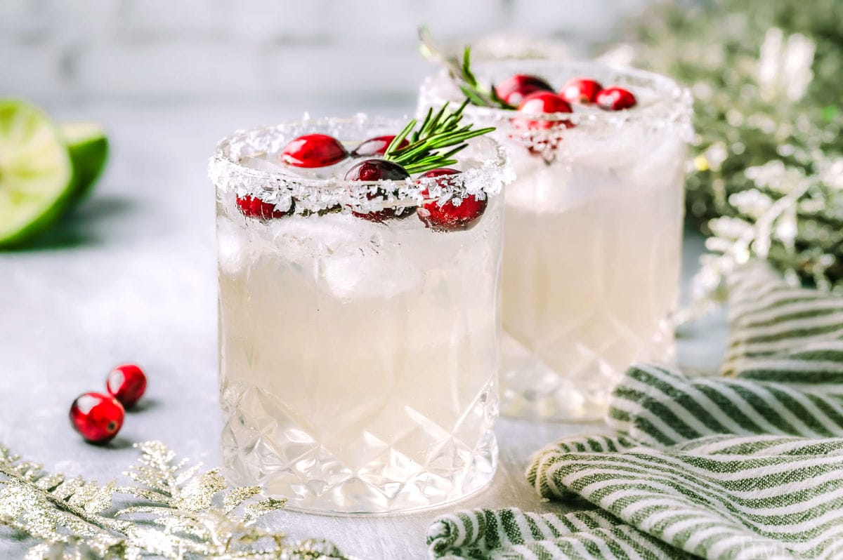 White Christmas Margarita recipe served in two glasses and topped with rosemary and cranberries. Margaritas are sitting next to a kitchen towel  and lime wedges and cranberries can be seen in the background.