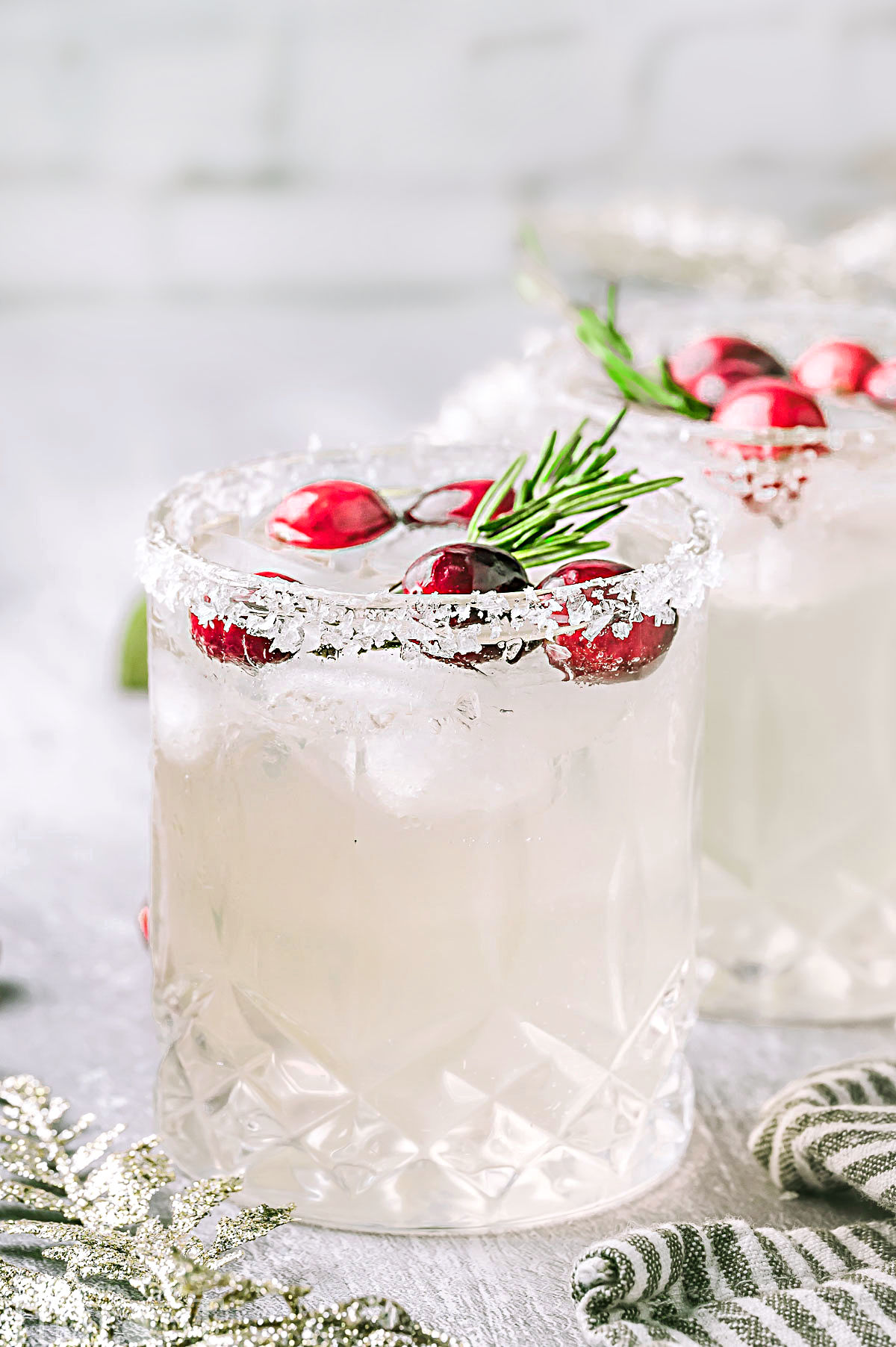 Two white christmas margaritas pictured on a white surface topped with cranberries and rosemary garnishes.