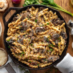 Top down view of cast iron skillet filled with mushroom pasta and finished with fresh parsley. Skillet is sitting on wood surface with mushrooms, shallots and herbs next to it.