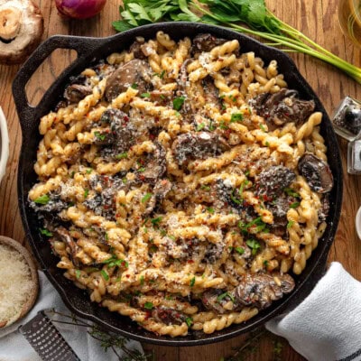 Top down view of cast iron skillet filled with mushroom pasta and finished with fresh parsley. Skillet is sitting on wood surface with mushrooms, shallots and herbs next to it.