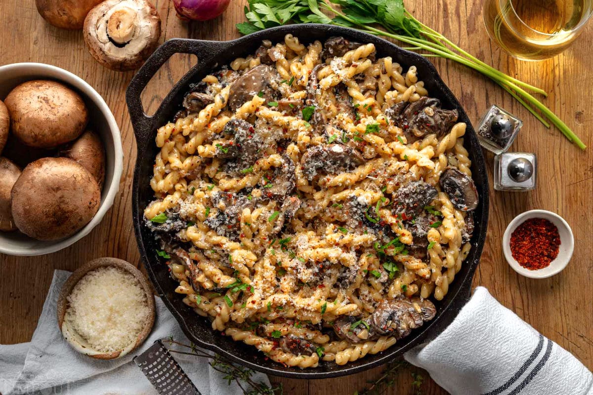 Top down view of cast iron skillet filled with mushroom pasta and finished with fresh parsley. Skillet is sitting on wood surface with mushrooms, shallots and herbs next to it.