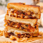 Shot of three halves of a french onion grilled cheese sandwich stacked on a wood cutting board with french fried onions surrounding it.