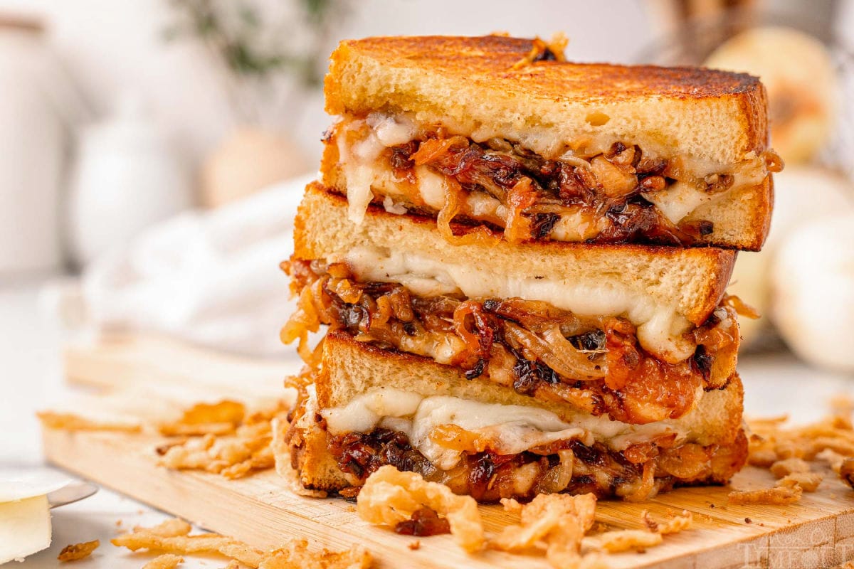 Wide shot of three halves of a french onion grilled cheese sandwich stacked on a wood cutting board with french fried onions surrounding it.