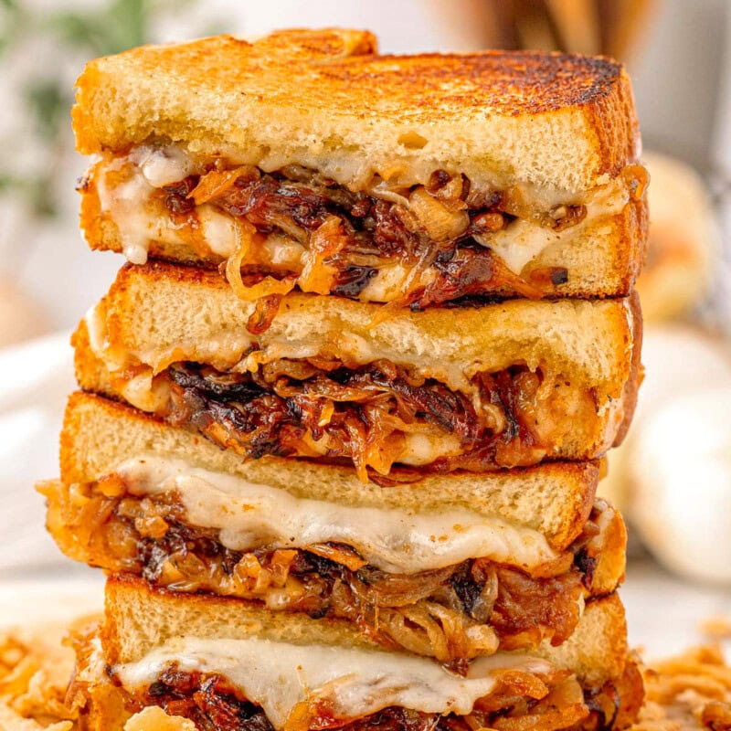 Tall stack of french onion soup grilled cheese cut in half on wood cutting board. Golden brown bread and gooey cheese can be seen along with the caramelized onion filling in the sandwiches.