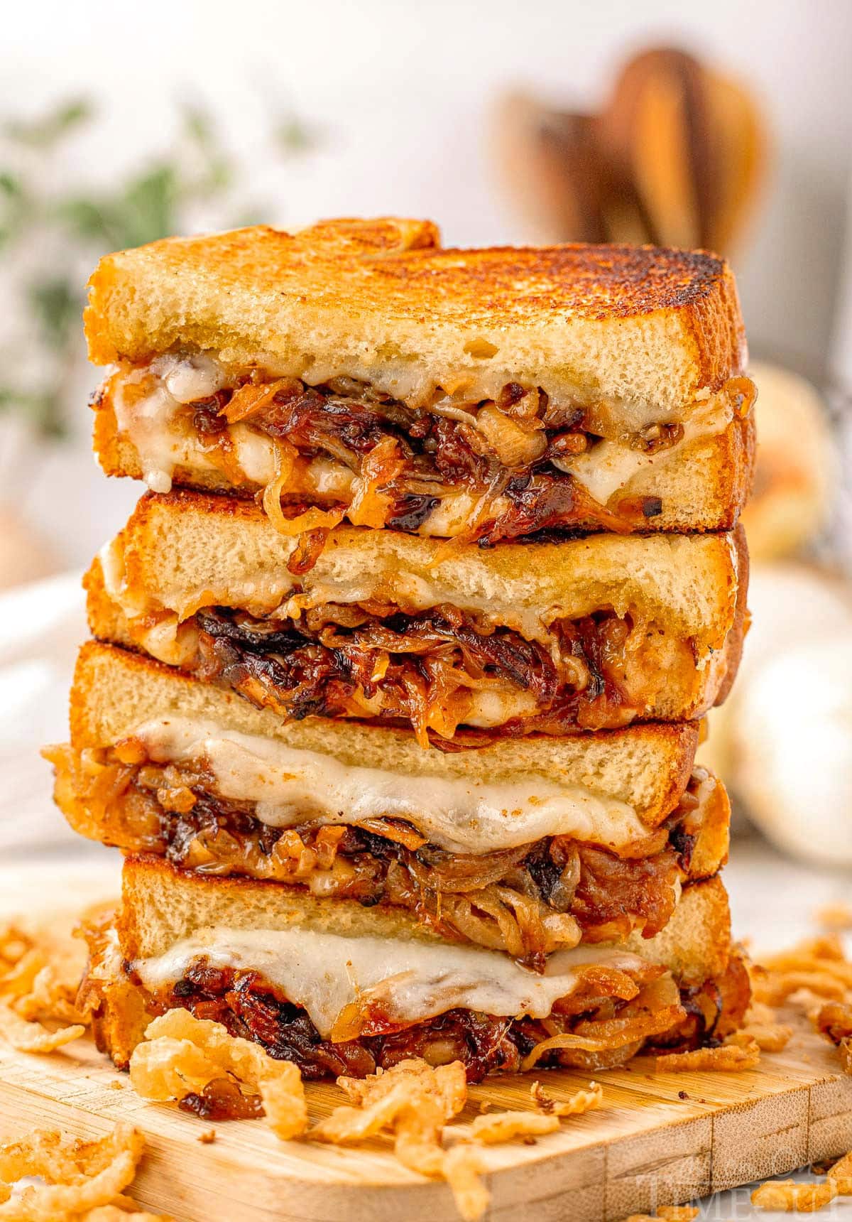 Tall stack of french onion soup grilled cheese cut in half on wood cutting board. Golden brown bread and gooey cheese can be seen along with the caramelized onion filling in the sandwiches.