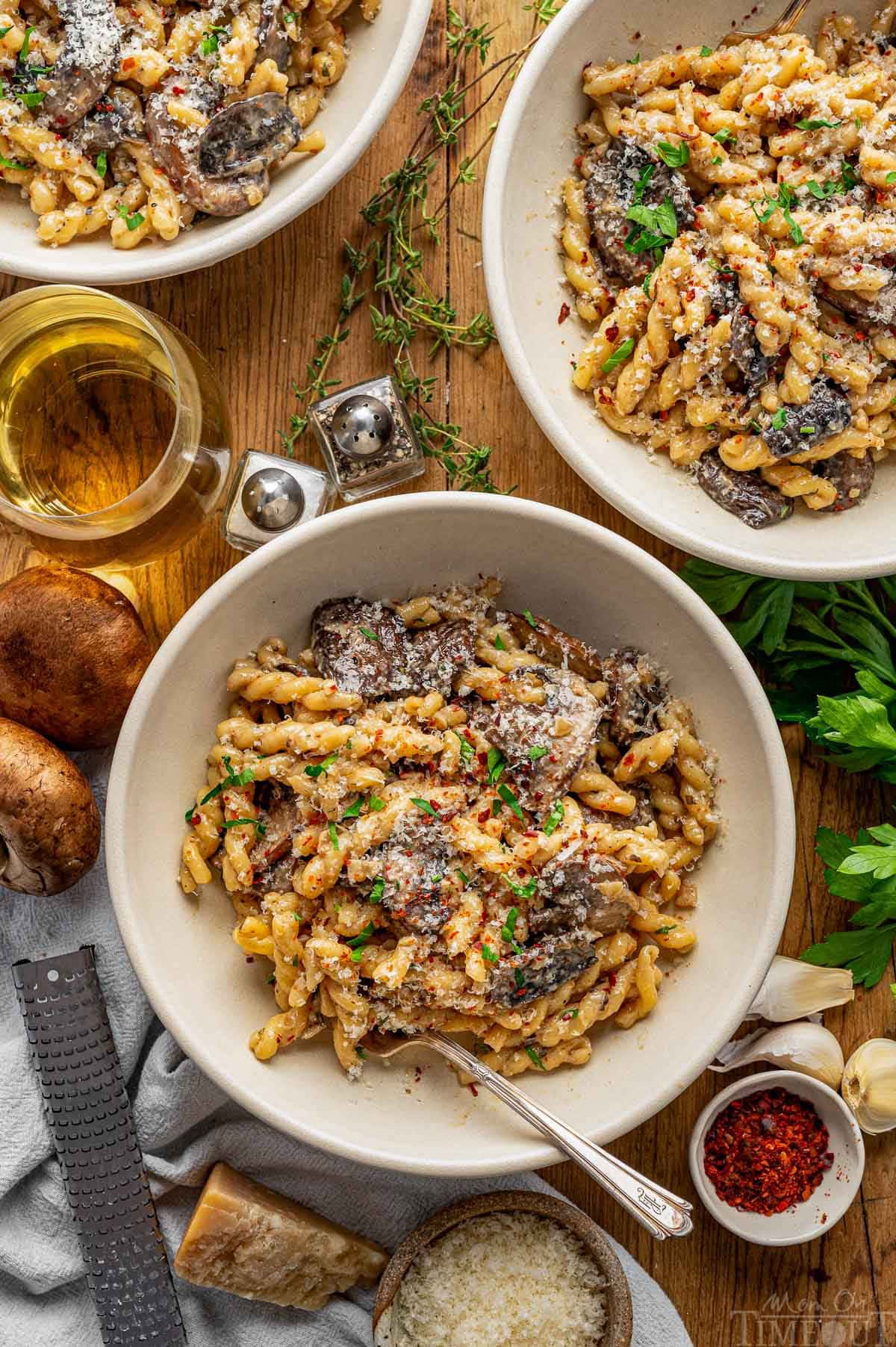 Top down shot of three bowls filled with a creamy mushroom pasta recipe. The bowls are on a wood surface next to a glass of wine, mushrooms, Parmesan and herbs.