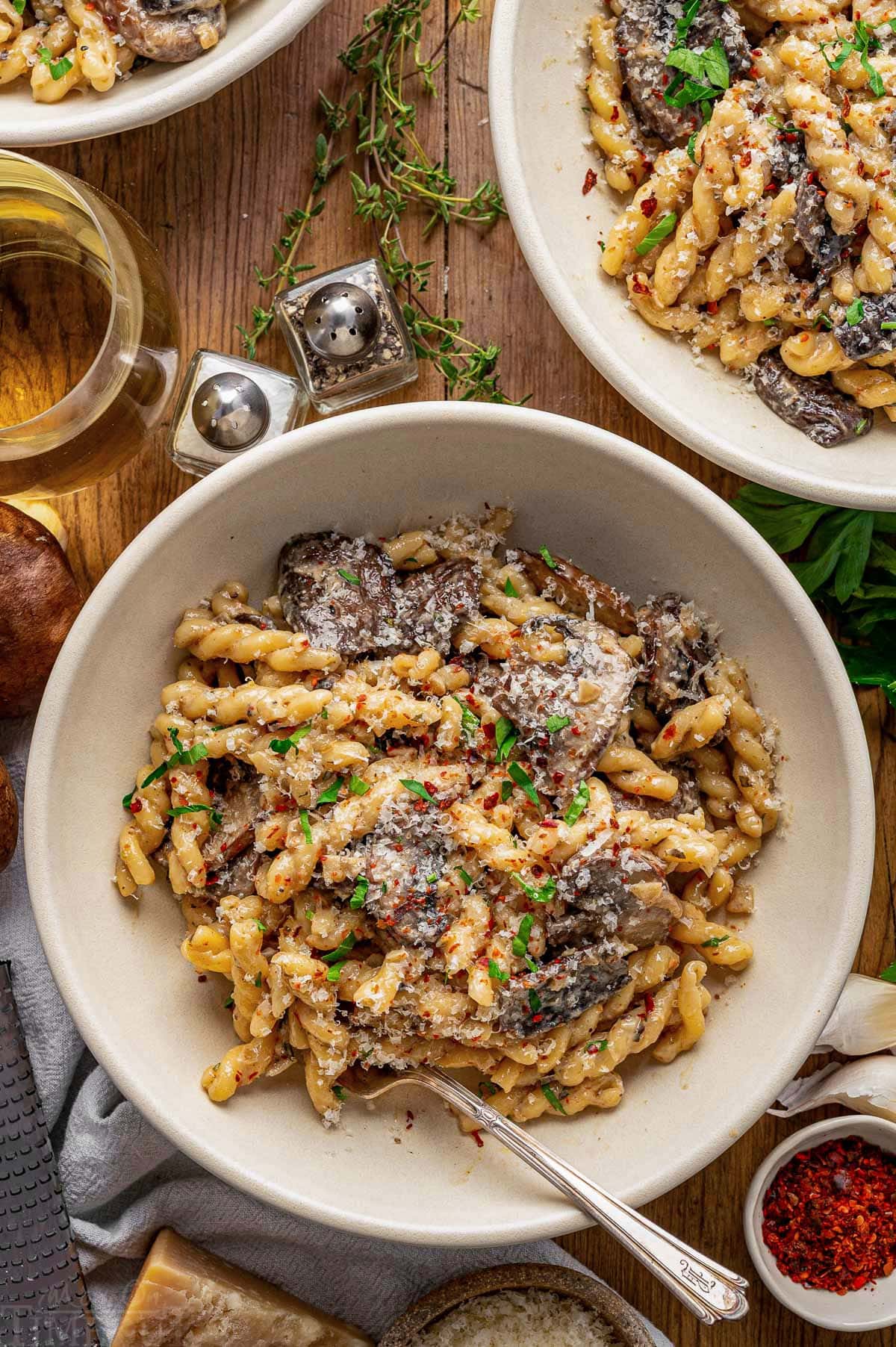 Top down shot of a white bowl filled with a mushroom pasta recipe. The bowl is on a wood surface next to a glass of wine, two more bowls, Parmesan and herbs.