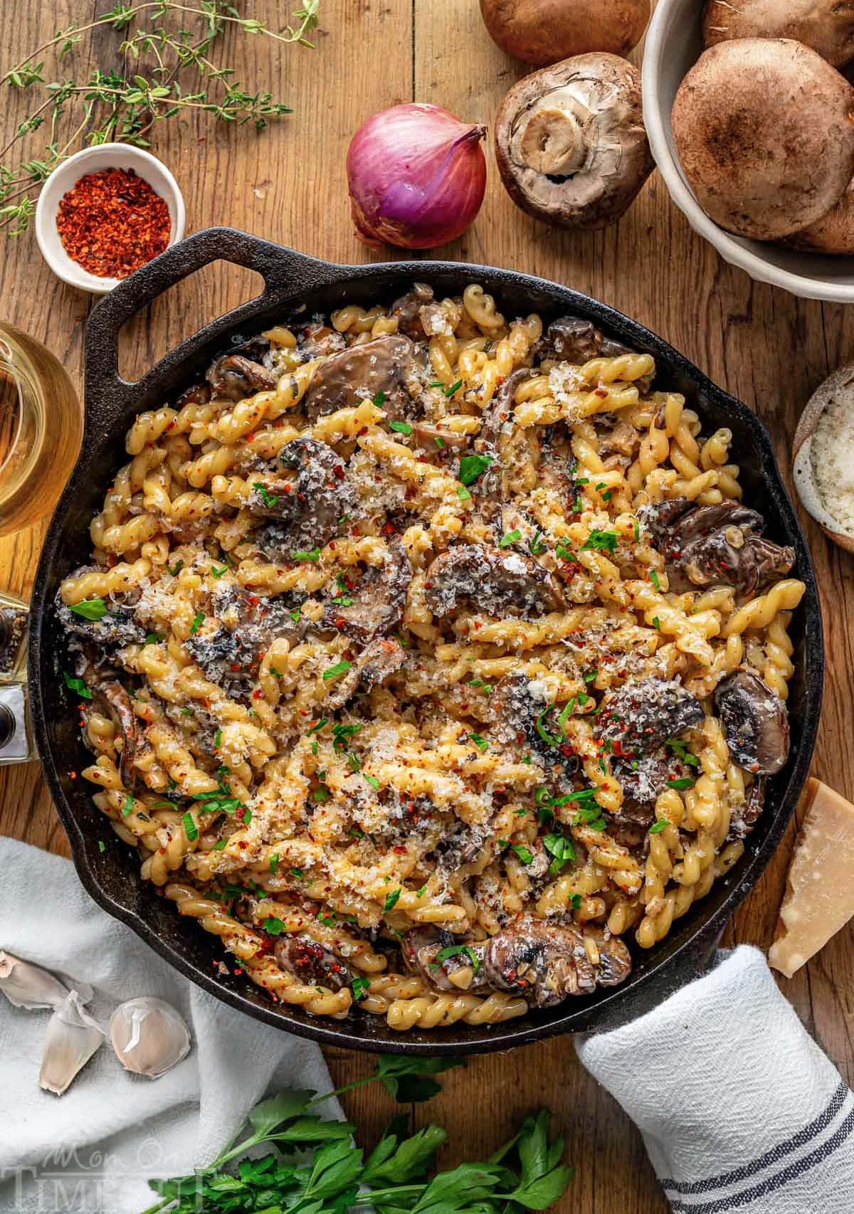 Top down view of cast iron skillet filled with mushroom pasta and finished with fresh parsley. Skillet is sitting on wood surface with mushrooms, shallots and herbs next to it.