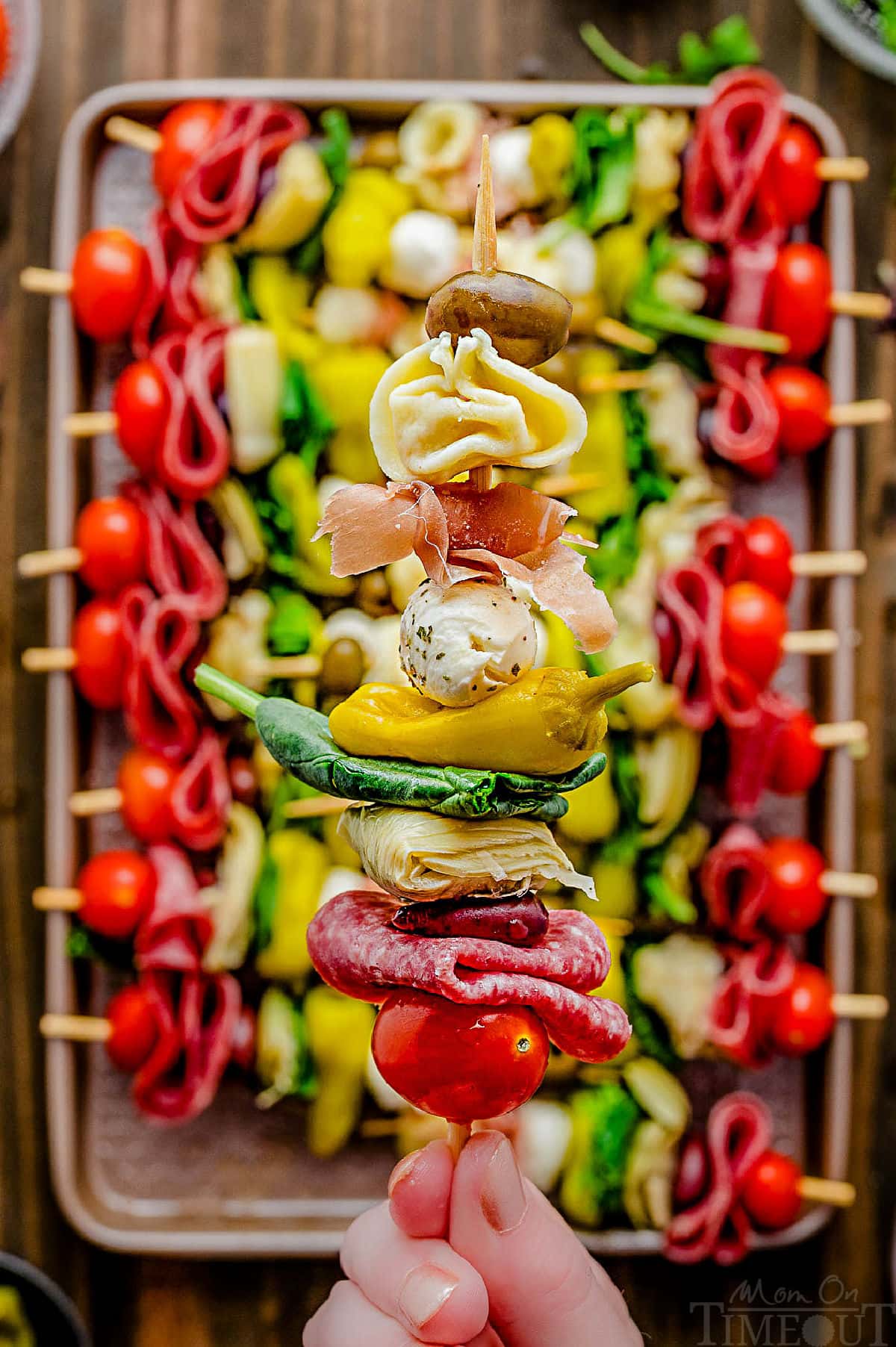 Single antipasto skewer being held over an entire sheet tray filled with more skewers.