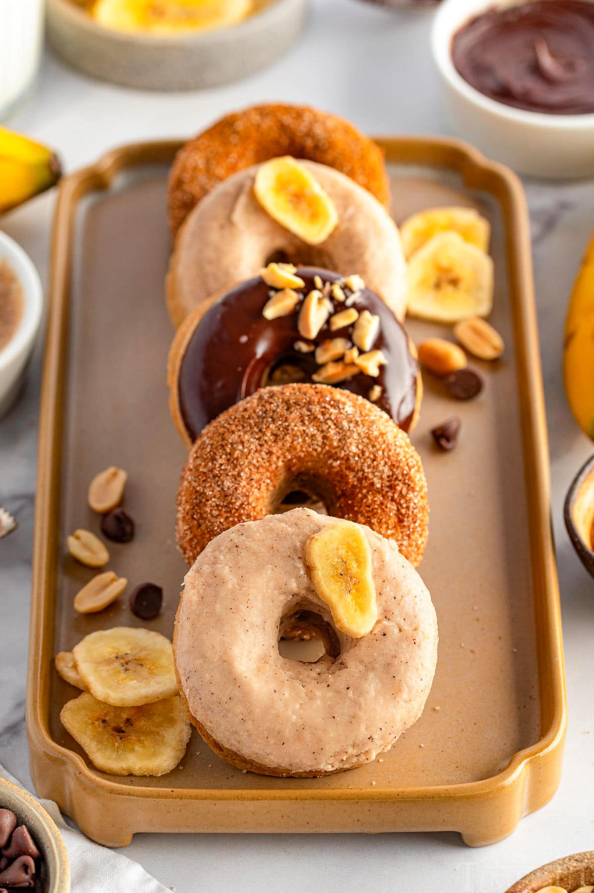 Banana Donuts leaning against each other on a pretty decorative tray. Toppings are on the tray next to the donuts.