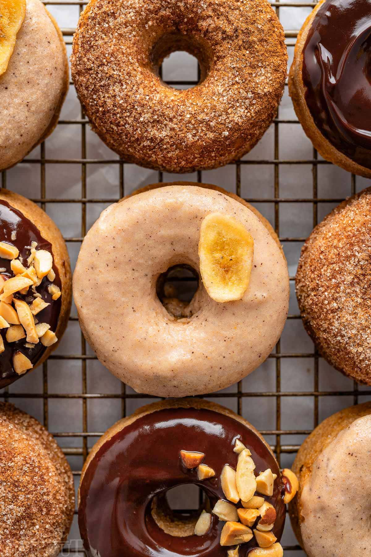 A top down view of banana donuts with various toppings. The donuts are on a metal cooling rack.