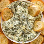 Crockpot spinach artichoke dip in a white bowl surrounded by crostini.