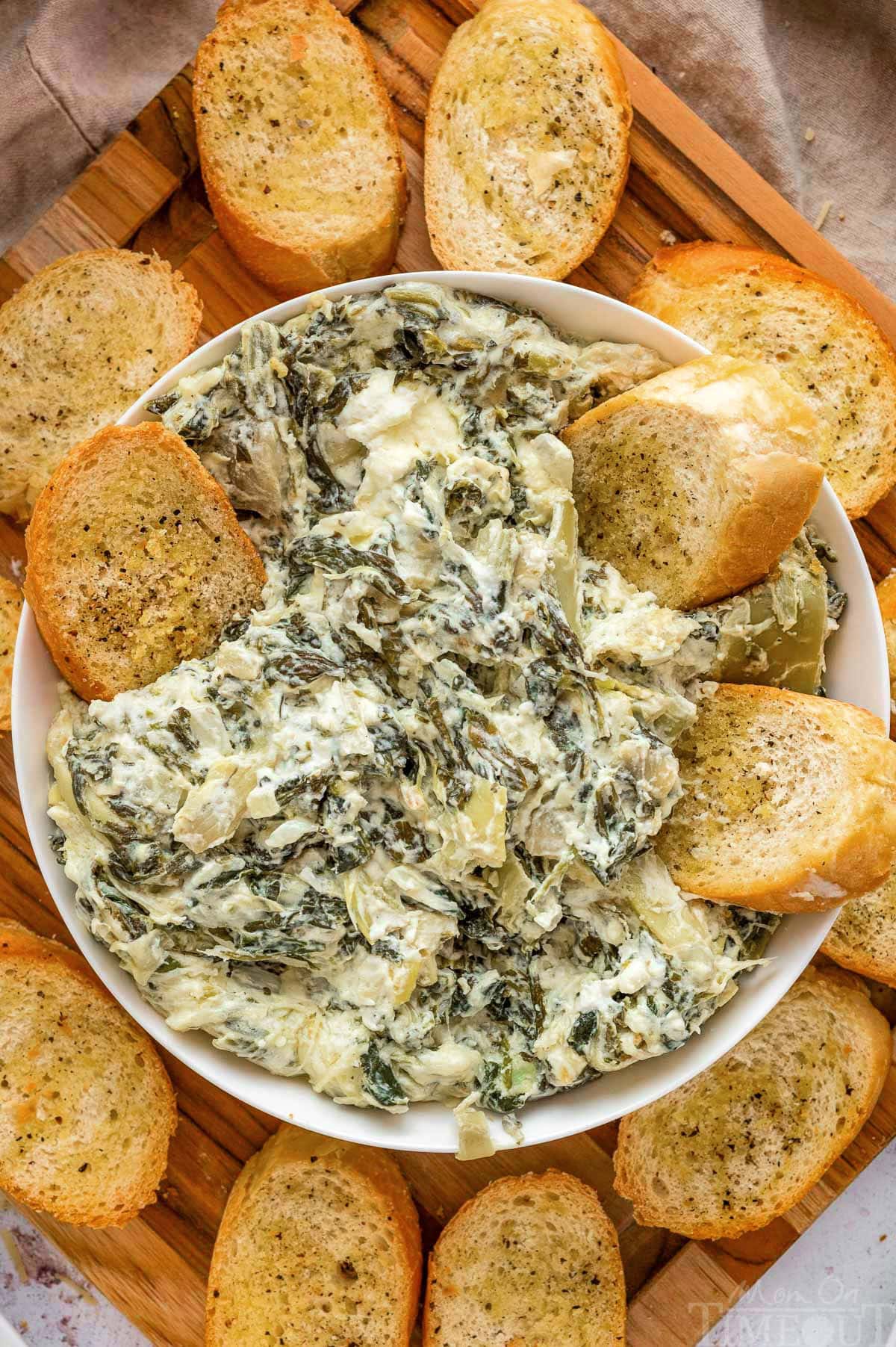 Crockpot spinach artichoke dip in a white bowl surrounded by crostini.