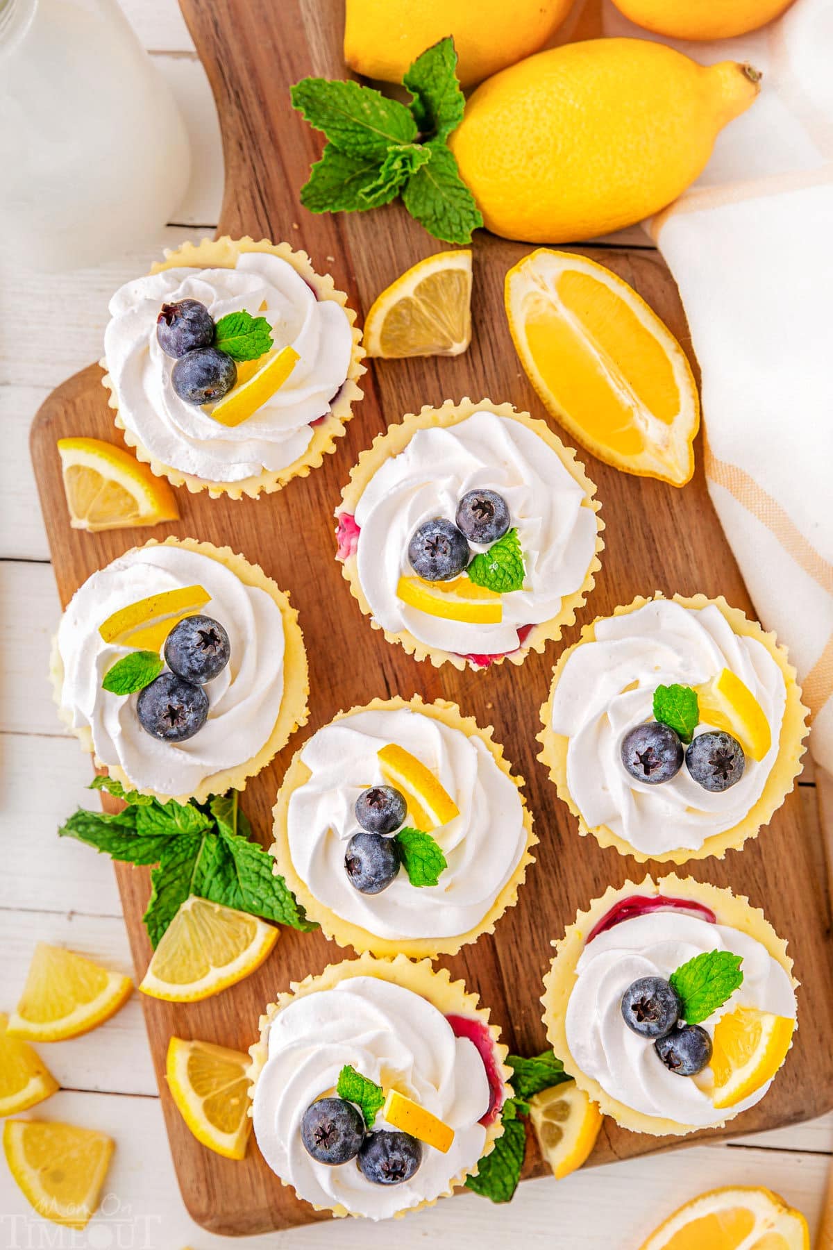 A top down view of lemon blueberry mini cheesecakes on a wood cutting board. The cutting board also has mint leaves and lemon slice garnishes on it.