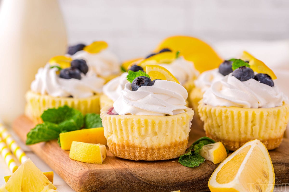 A side view of lemon blueberry mini cheesecakes on a wood cutting board. Garnishes are scattered around the cheesecakes.