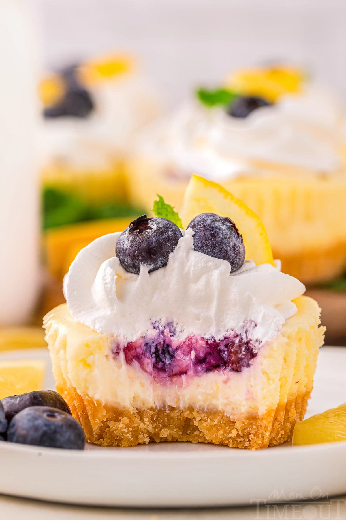 A close up view of a lemon blueberry mini cheesecake with a bite taken out of it. The cheesecake is on a white plate next to garnishes.