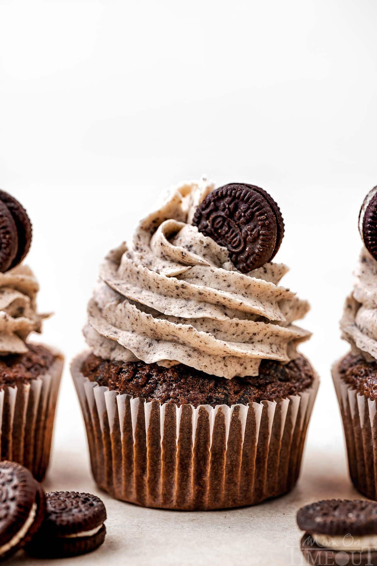 Three cookies and cream cupcakes lined up and topped with oreo frosting and a mini oreo on a white background.