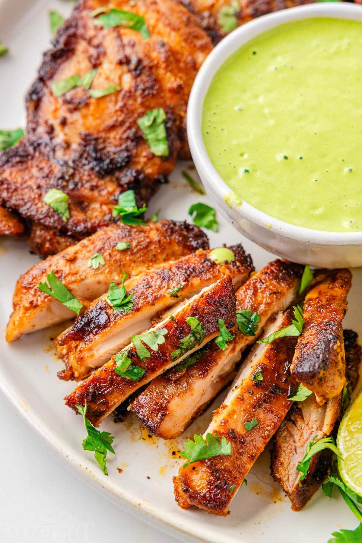 Sliced Peruvian chicken on a white serving plate next to a bowl with aji verde.