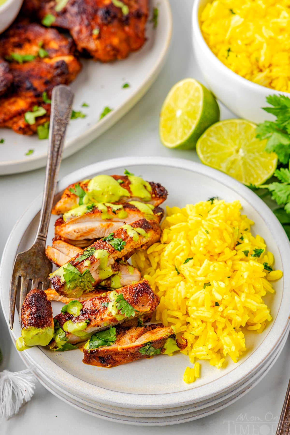 A top down view of sliced Peruvian Chicken next to a side of rice on a white plate with a fork holding a piece. Extra chicken and rice are on serving plates in the background.